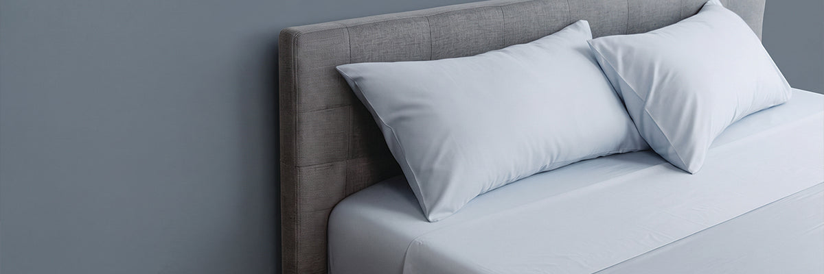 A close-up of a bed with white pillows and white bedding, set against a gray upholstered headboard and a muted blue-gray wall.