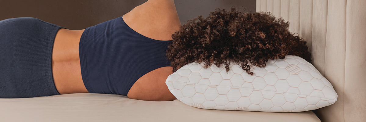 A person lying on a bed with their back to the camera, wearing a navy blue sports bra and shorts, resting on a white pillow with a honeycomb pattern.